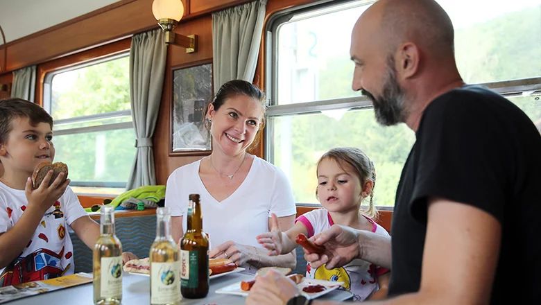 Eine vierköpfige Familie sitzt an einem Tisch in einem nostalgischen Zugabteil. Auf dem Tisch stehen Getränkeflaschen, Teller mit Würstchen und Senf sowie Brotscheiben.