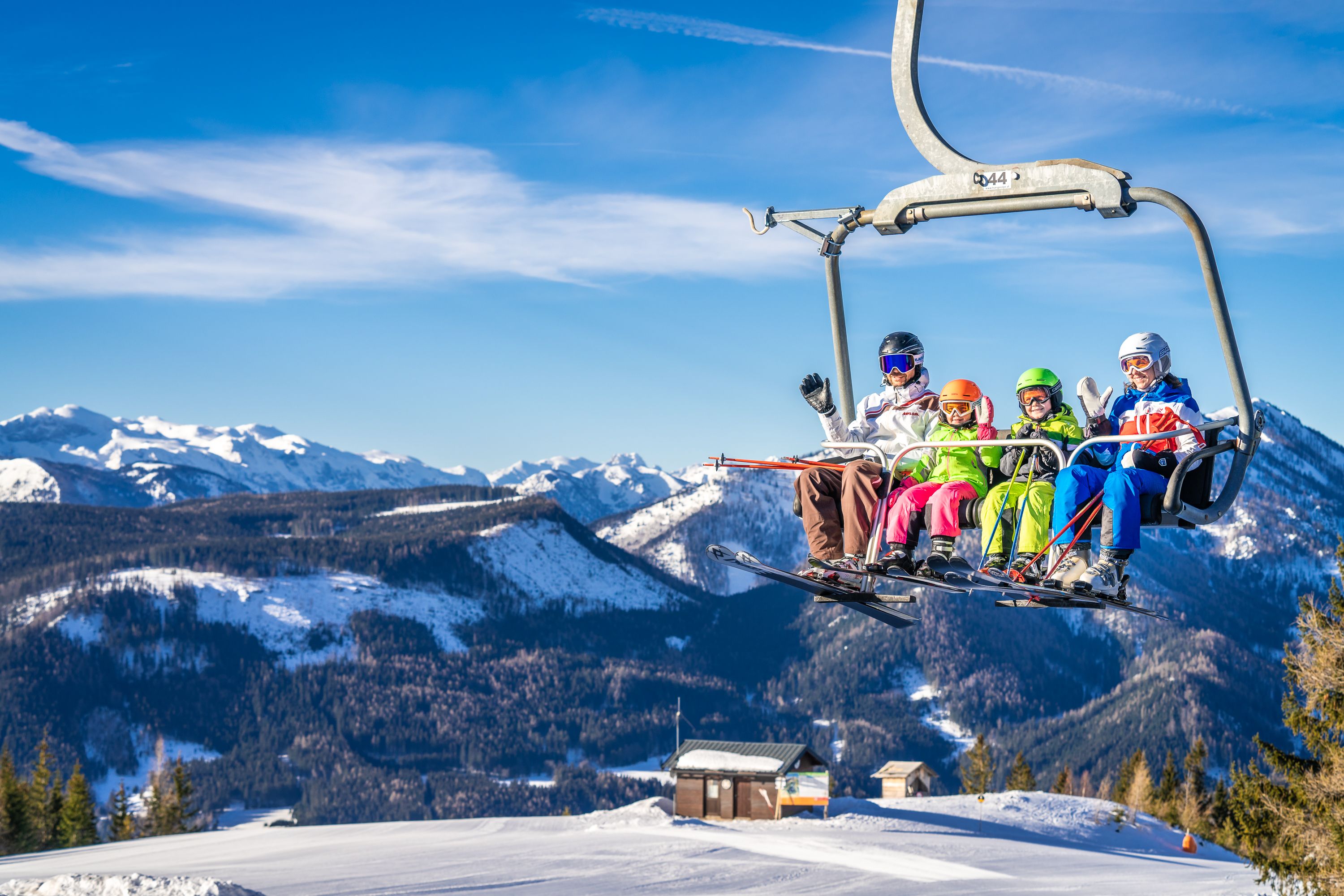 Zwei Erwachsene und zwei Kinder fahren mit Ski und Snowboards im Sessellift vor verschneiter Bergkulisse.