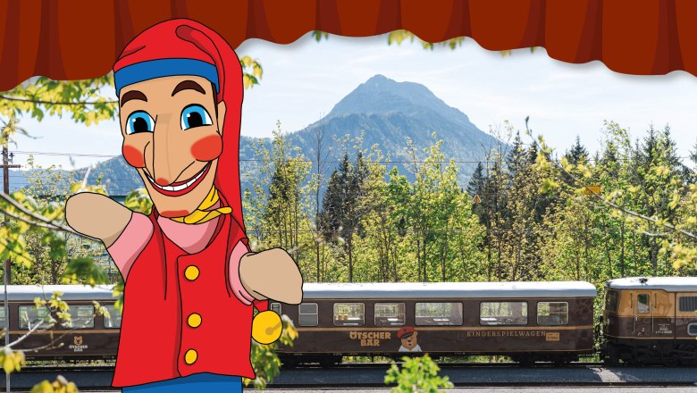 Eine lächelnde Kasperlfigur vor dem Ötscherbärzug. Im Hintergrund sind Bäume und ein Berg zu sehen.