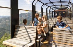 Eine Familie sitzt auf Holzbauteilen in einem offenen Aussichtswagen eines Zuges und schaut auf eine bergige Waldlandschaft unter sonnigem Himmel.