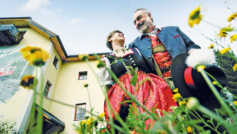 Ein Paar in traditioneller Tracht steht vor einem gelben Geb&auml;ude, umgeben von Blumen, und l&auml;chelt sich an.