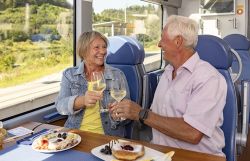 Eine ältere Frau und ein älterer Mann stoßen im Zug mit Weißweingläsern über einem gedeckten Tisch an.