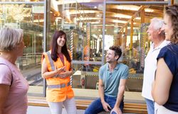 Fünf Personen in einer Eisenbahnwerkstatt, eine Frau trägt eine orange Warnweste und spricht, die anderen hören zu und lächeln.