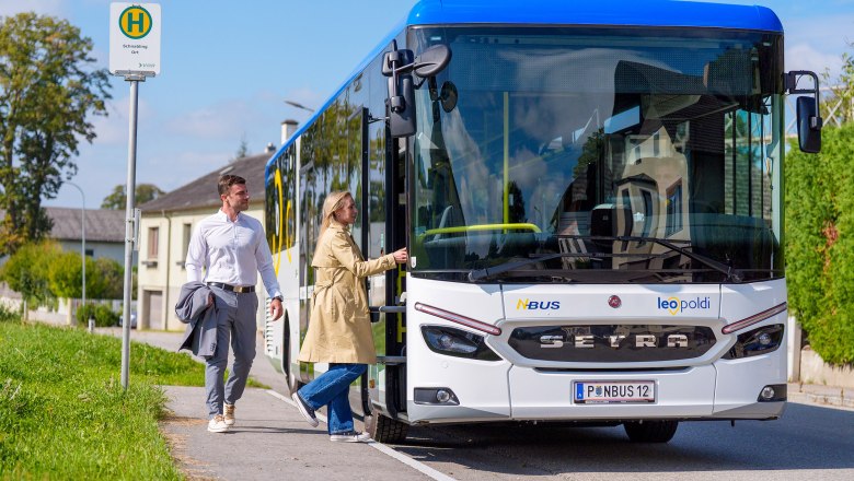 Blau weißer Linienbus in ländlicher Umgebung.  2 Personen steigen in den Bus ein.