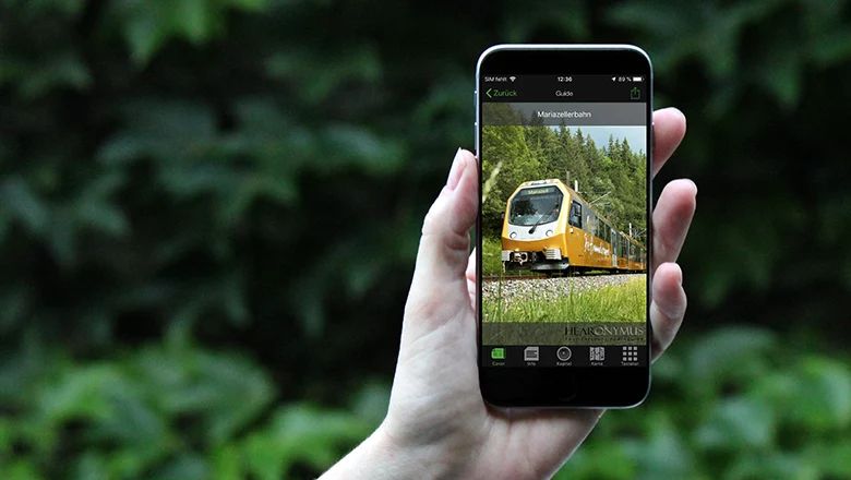 Hand hält Smartphone mit Bild eines gelben Zuges auf dem Bildschirm und der Überschrift 'Mariazellerbahn'