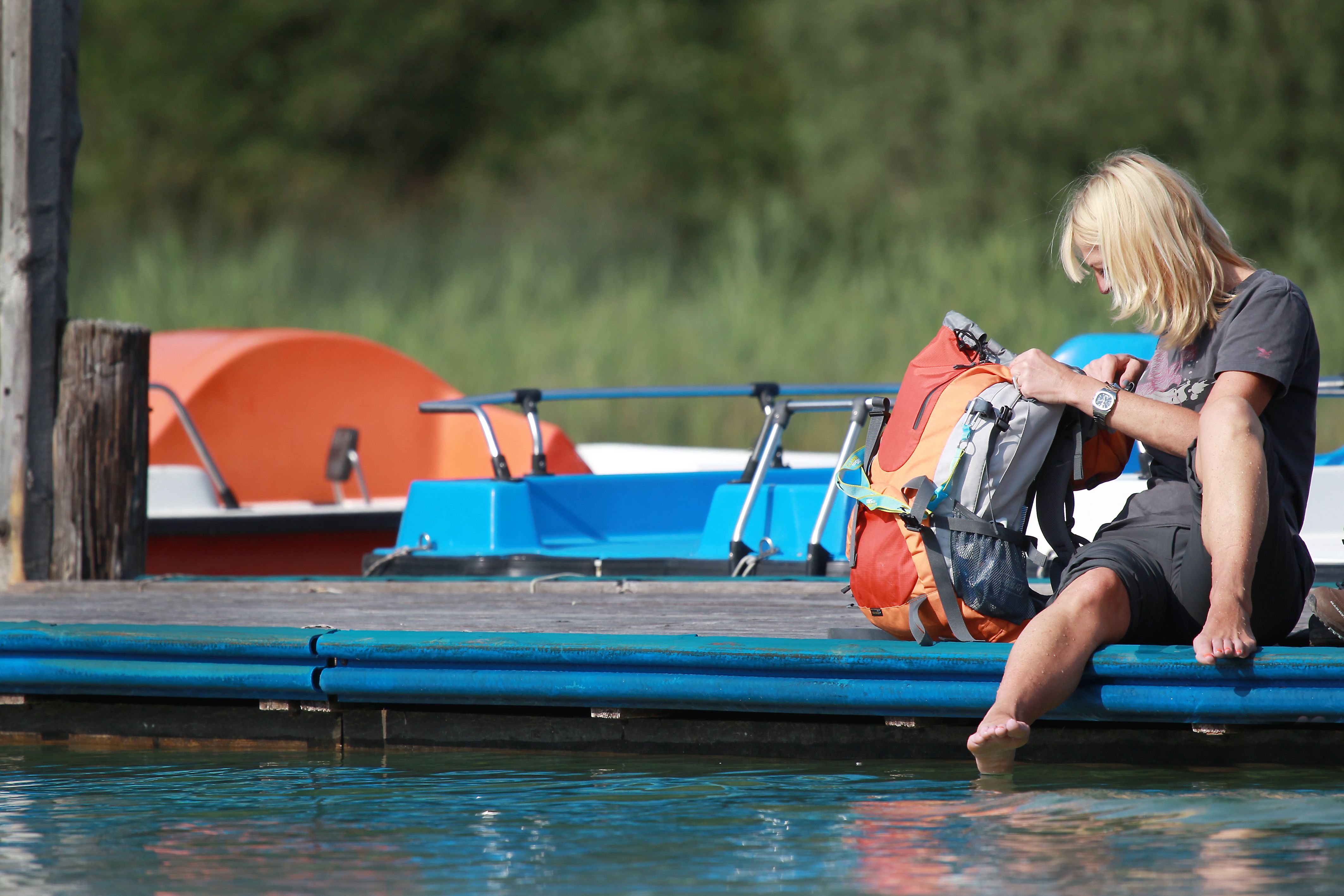 Person sitzt auf einem Steg am Erlaufsee mit den Füßen im Wasser, neben einem Rucksack. Im Hintergrund sind Tretboote zu sehen.
