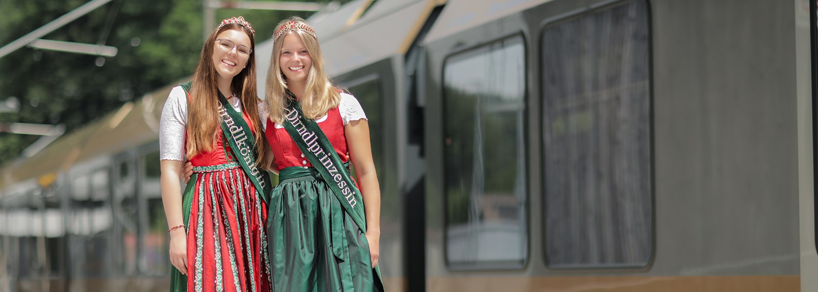 Zwei Frauen in traditioneller Tracht mit roten und grünen Kleidern und Schärpen, stehen vor einem Zug.
