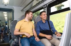 Zwei Männer sitzen nebeneinander auf Passagiersitzen in einem modernen Zugabteil, unterhalten sich und lächeln. Durch das große Fenster ist eine grüne Hügellandschaft zu sehen.