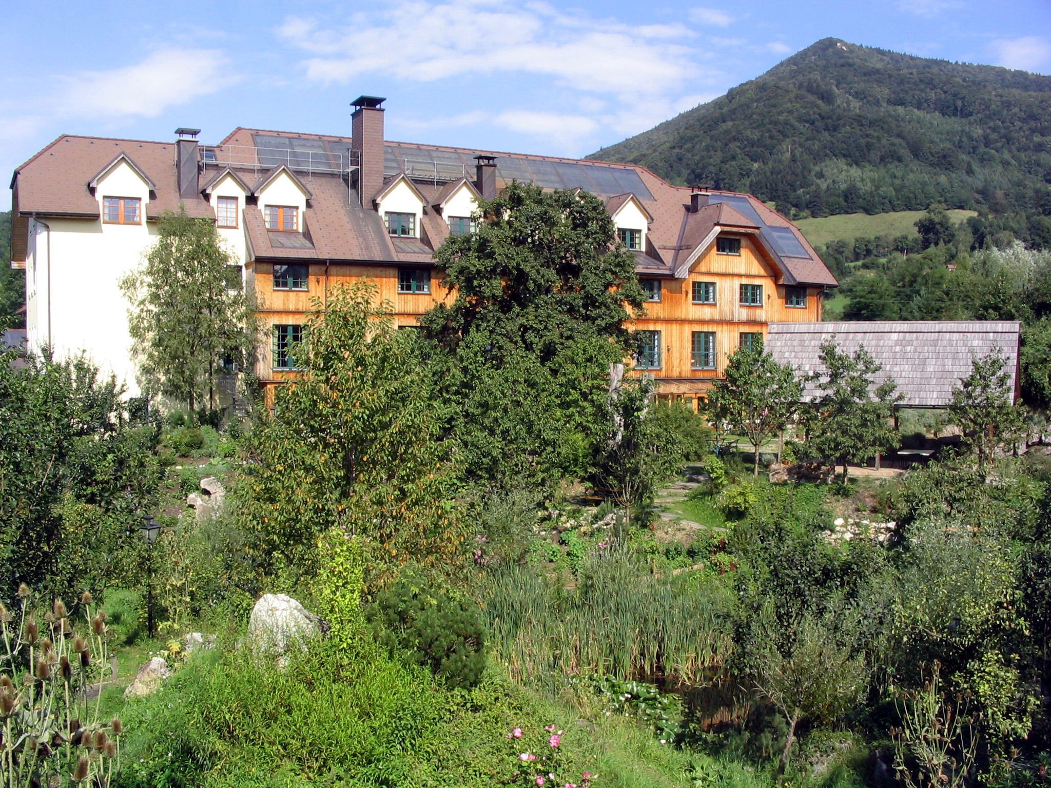 Ein großes Gebäude mit Holzfassade, umgeben von üppigem Grün und einem Berg im Hintergrund.
