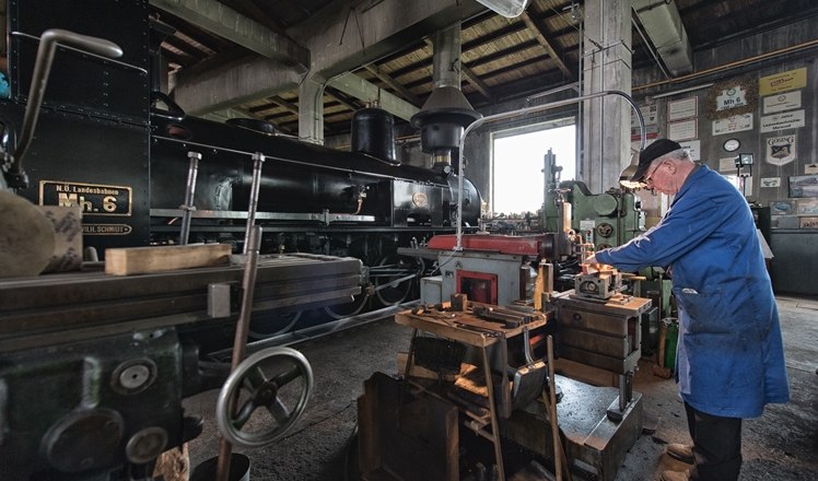 Ein Mann arbeitet in einer Werkstatt an einer Maschine neben einer alten Dampflokomotive.
