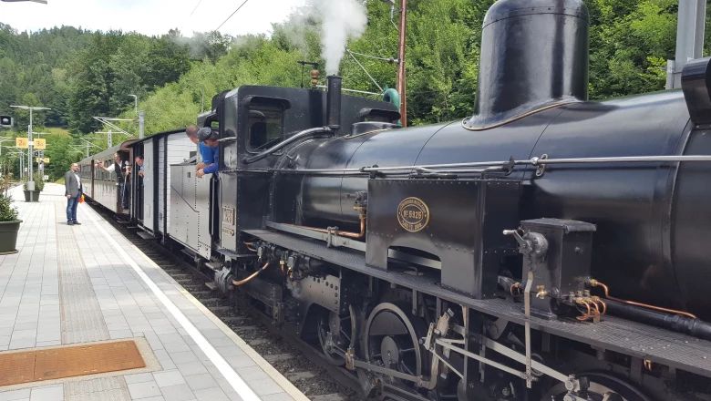 Eine schwarze Dampflokomotive steht mit angehängten Waggons an einem gepflasterten Bahnsteig. Personen schauen aus den Zugtüren und Fenstern heraus.