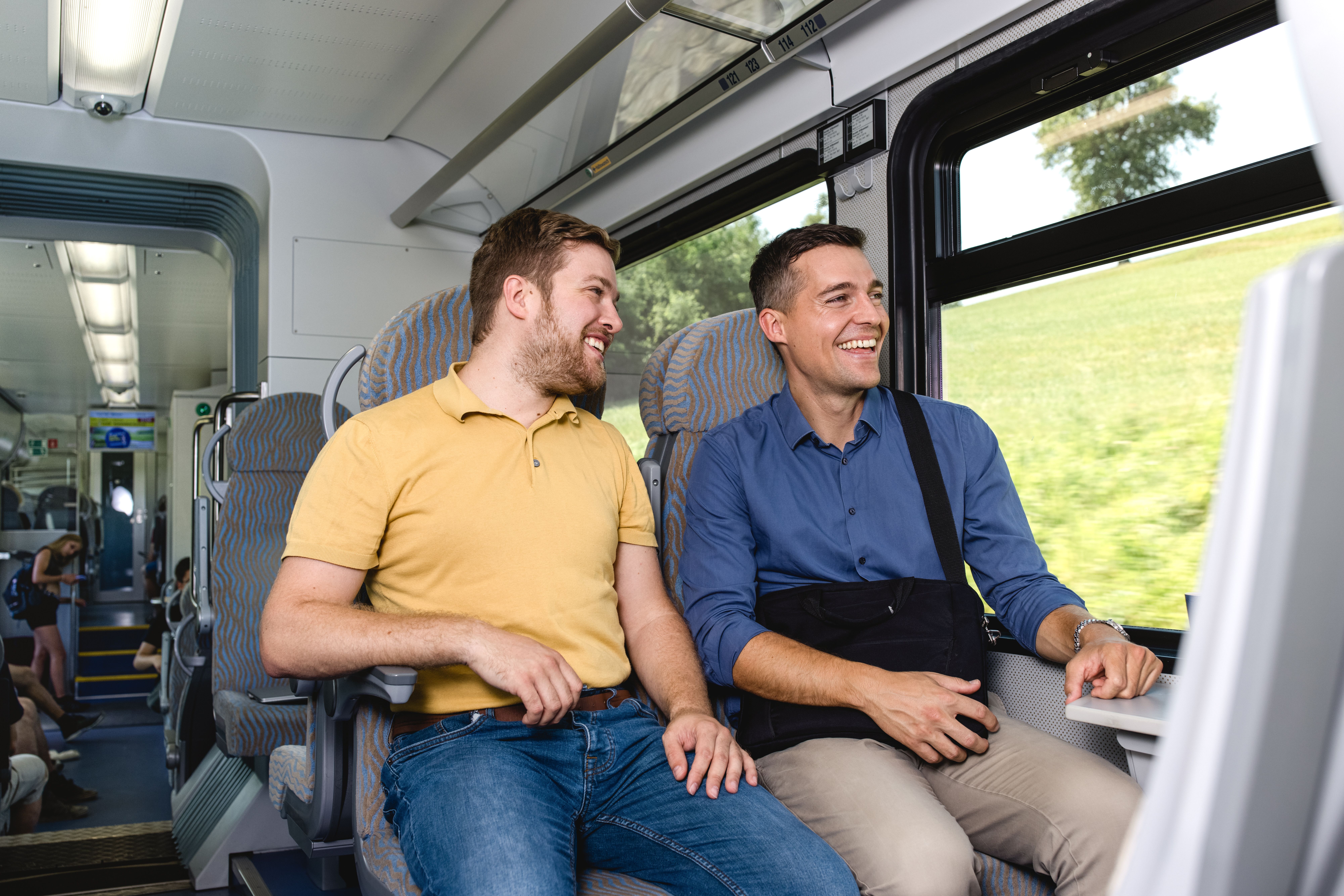 Zwei Männer sitzen nebeneinander auf Passagiersitzen in einem modernen Zugabteil, unterhalten sich und lächeln. Durch das große Fenster ist eine grüne Hügellandschaft zu sehen.