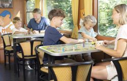 Kinder und Erwachsene sitzen im Spielewagen eines Zuges an Tischen und spielen Gesellschaftsspiele mit Spielfiguren und Würfeln. An der Wand befindet sich ein Bärenmotiv.
