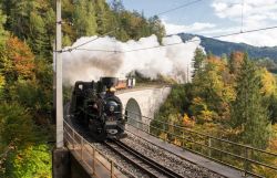 Ein Dampfzug fährt über ein steinernes Viadukt. Weiße Dampfwolken steigen in den Himmel auf, umgeben von herbstlich gefärbten Bäumen und Hügeln.