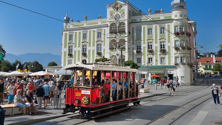 Historische rot-gelbe Straßenbahn fährt über einen belebten Stadtplatz. Im Hintergrund steht ein reich verziertes grün-weißes Gebäude unter blauem Himmel.